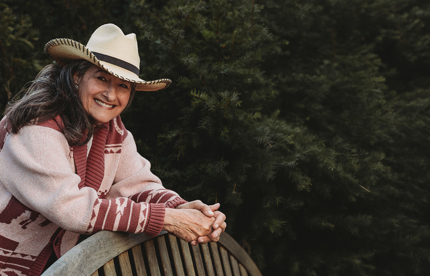 An older woman wearing a cowboy hat and a patterned cardigan leans on a curved wooden bench, smiling warmly, with green foliage in the background.